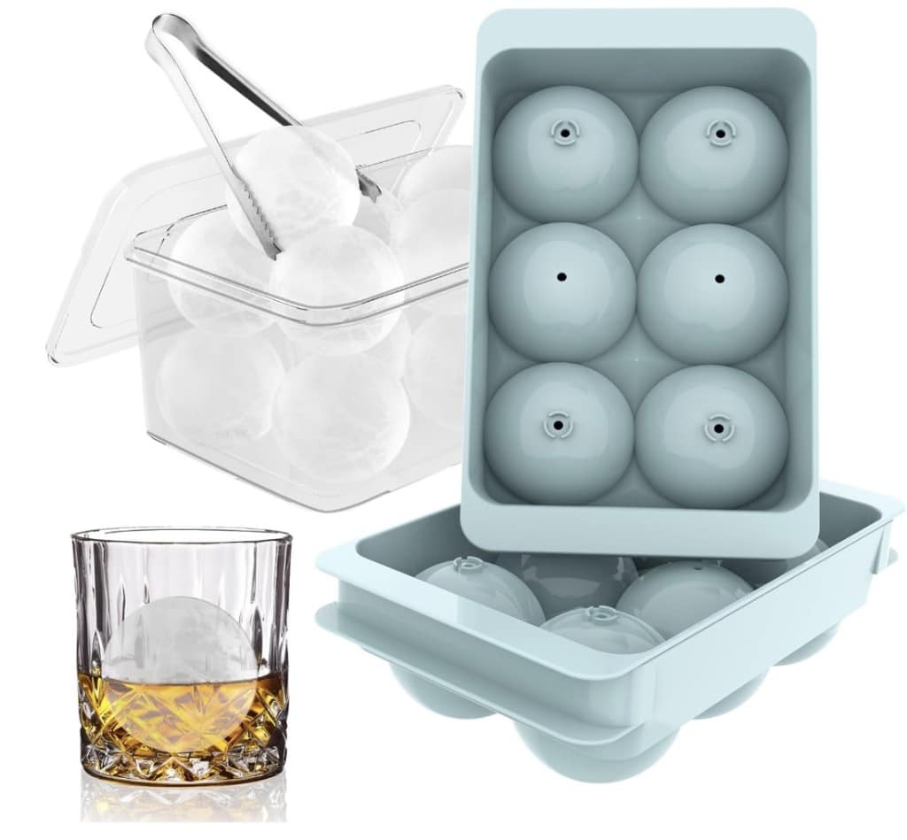 A six-cavity silicone mold for making round ice spheres sits open. Beside it, a clear container holds ice balls with tongs resting on top. In the foreground, a glass of whiskey features one ice sphere.